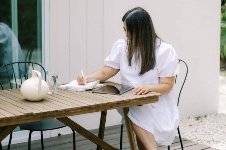 A woman seated at a table, engrossed in writing in a notebook, seeking ways to simplify her life.