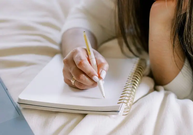 A woman sitting on a bed, writing in a notebook. She is focused and determined, using her time wisely to organize her life effectively.- organize your life
