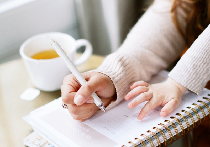 A mom journaling with with a cup of drink and her babies hands also on the desk