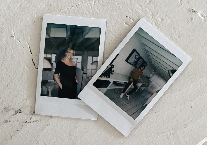 This image shows two Polaroid-style photographs placed on a textured, neutral-colored surface. The left photo features a woman in a black dress posing indoors with a soft smile, while the right photo shows a man standing casually in what appears to be a modern, minimalist room with white floors and slanted ceilings. Both images evoke a cozy, personal, and nostalgic vibe.