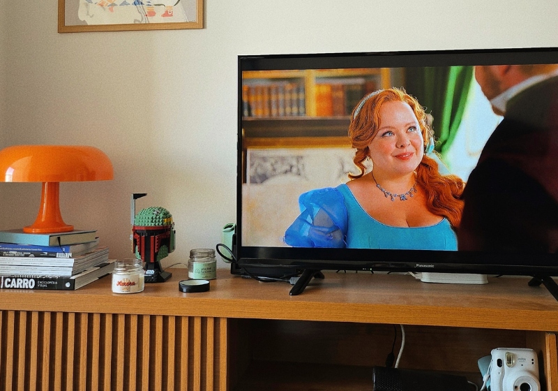 A living room setup with a TV displaying a scene from Netflix Bridgerton drama. A wooden stand holds books, candles, a model, and an orange lamp.