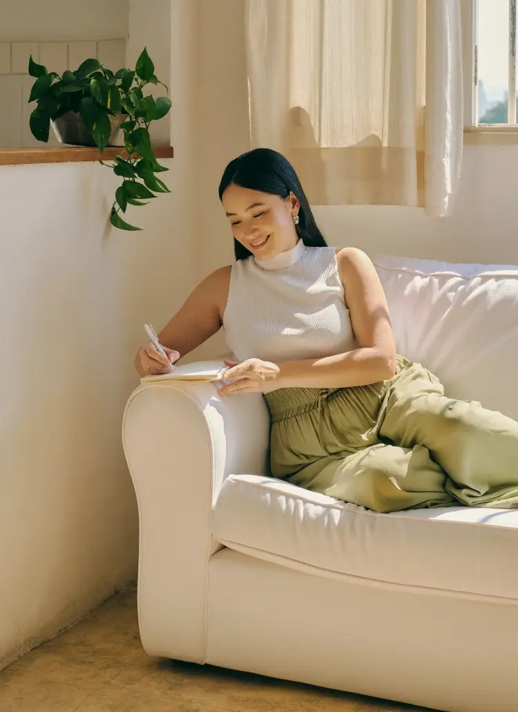 A woman sitting on a white couch, smiling as she writes in a notebook, symbolizing calm reflection and positivity—perfect for journaling affirmations for protection