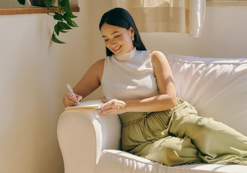 A woman sitting on a white couch, smiling as she writes in a notebook, symbolizing calm reflection and positivity—perfect for journaling affirmations for protection