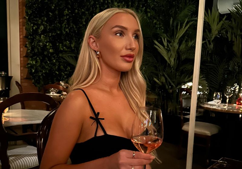 The image shows a woman with long blonde hair sitting at a table outdoors. She is wearing a black dress and is holding a glass of rosé wine. The background features lush greenery, suggesting an outdoor dining setting