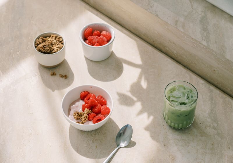  a healthy breakfast or snack setup. It shows bowls of granola and fresh strawberries, as well as a green drink that appears to be some kind of smoothie or juice. The items are arranged on a light-colored surface, creating a clean and minimalist presentation. The natural lighting from the window creates interesting shadows and highlights the colors of the food item—get your life together.