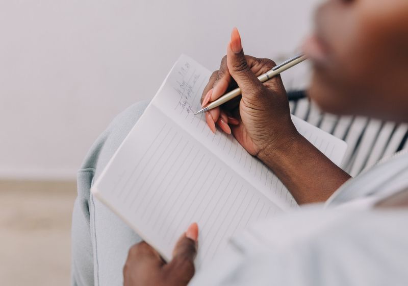 A woman writing in a notebook. The person is holding a pen and appears to be carefully writing or taking notes on the lined pages of the notebook. The setting is simple and focused on the act of journaling.