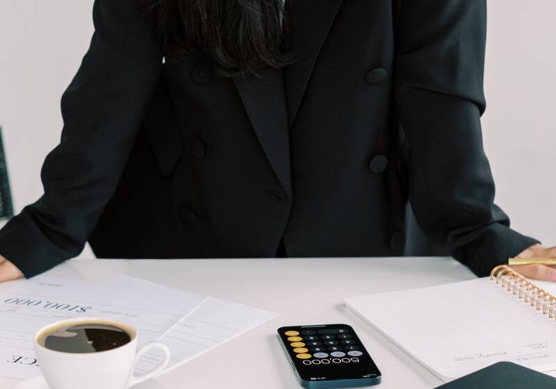 A woman in a black business suit standing at a desk, with a coffee mug and notebook visible.