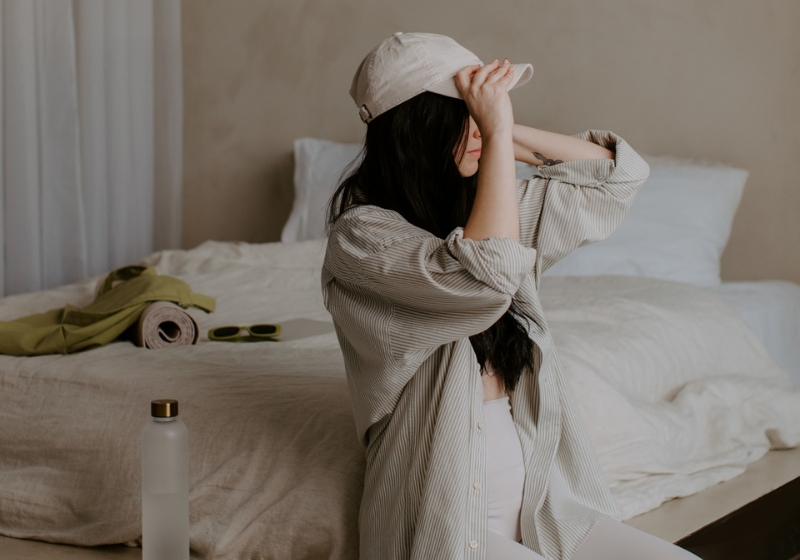 A woman sitting on the edge of a bed in a minimalistic room, adjusting a beige baseball cap. She is wearing a striped oversized shirt and white leggings, with a reusable water bottle and neatly folded items visible nearby, evoking a calm and organized atmosphere. Perfect for illustrating themes of mindfulness and getting life together.