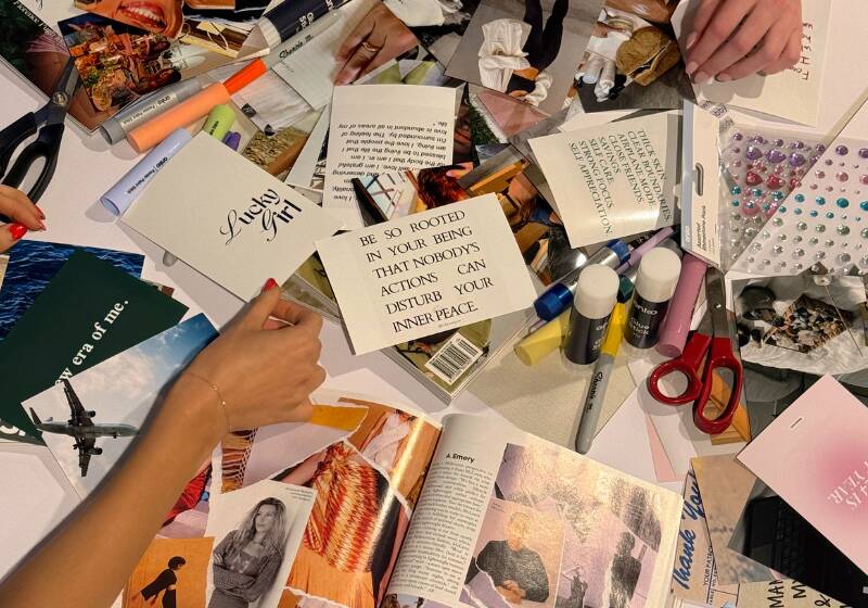 Overhead view of a craft workspace showing hands working on what appears to be a vision board or collage project. The surface is covered with magazine clippings, photographs, quotes (including one visible quote that reads 'BE SO ROOTED IN YOUR BEING THAT NOBODY'S ACTIONS DISTURB YOUR INNER PEACE'), markers, scissors, and decorative elements like adhesive gems. An open magazine is visible in the lower portion of the image