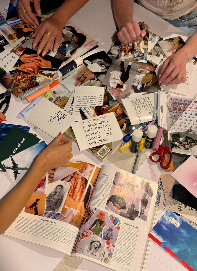 Overhead view of a craft workspace showing hands working on what appears to be a vision board or collage project. The surface is covered with magazine clippings, photographs, quotes (including one visible quote that reads 'BE SO ROOTED IN YOUR BEING THAT NOBODY'S ACTIONS DISTURB YOUR INNER PEACE'), markers, scissors, and decorative elements like adhesive gems. An open magazine is visible in the lower portion of the image