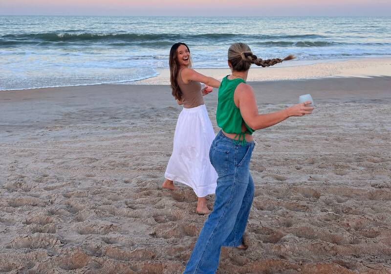 The image shows two young women enjoying a sunset at the beach. One woman is wearing a green top and the other is wearing a white skirt. They are walking in the sand and appear to be laughing and taking a photo together.