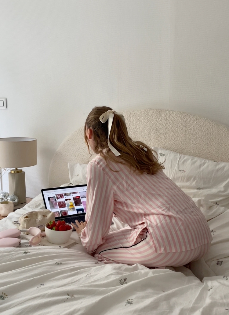 The image shows a woman sitting on a bed, surrounded by various items like a laptop, strawberries, and a lamp. She appears to be wearing a pink and white striped robe and has her hair tied up -a featured image iin a blog post about how to enjoy life fully.