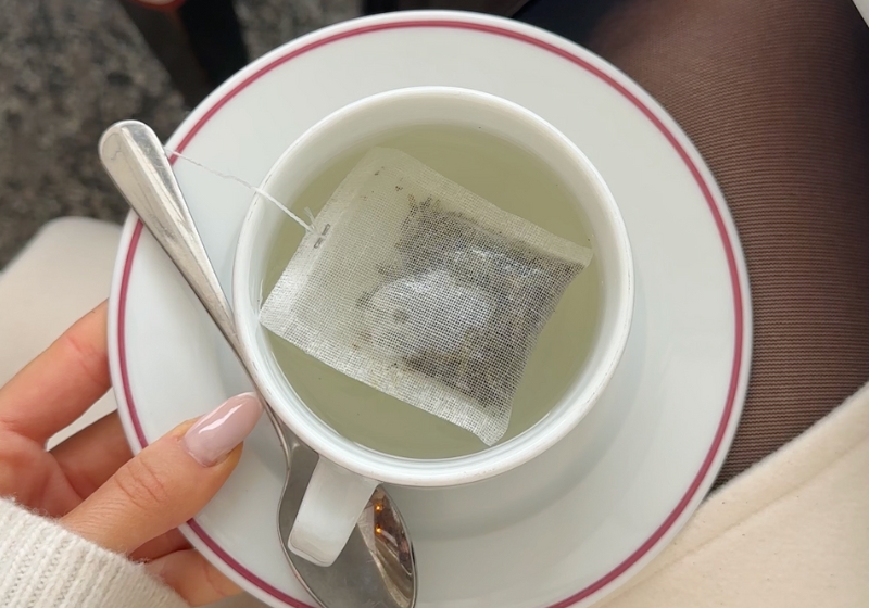 The image shows a cup of green tea with a tea bag in it. The tea appears to be steeping, and the cup is placed on a white and pink plate.