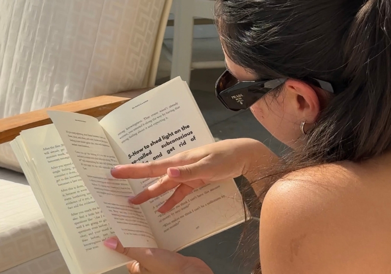 The image shows a woman reading a book while sitting on a bed. The person appears to be wearing dark clothing and sunglasses as they intently focus on the book. Copy