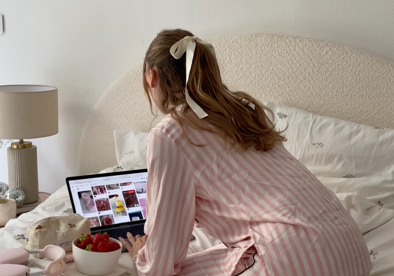 The image shows a woman sitting on a bed, surrounded by various items like a laptop, strawberries, and a lamp. She appears to be wearing a pink and white striped robe and has her hair tied up.