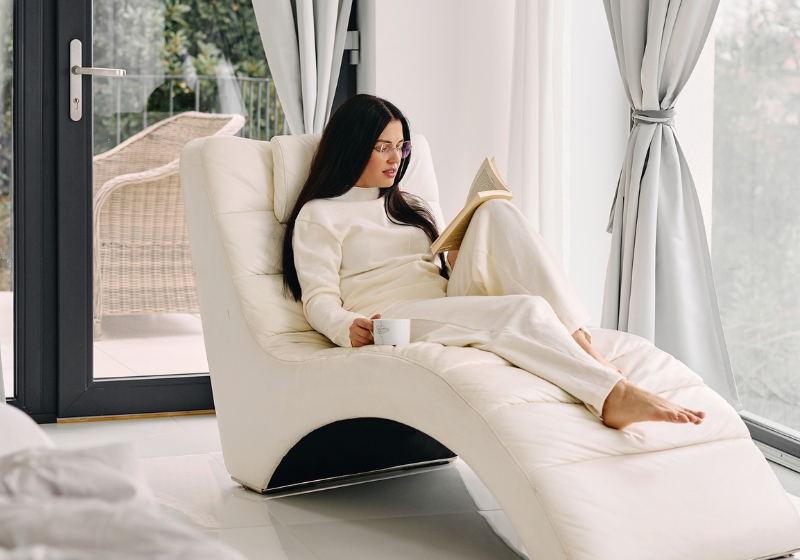  person relaxing on a white chaise lounge chair by large windows, reading a book and holding a coffee cup. They are wearing a cozy cream-colored sweater and matching loungewear, creating a serene monochromatic scene. A featured image in this blog post about Friday affirmations.