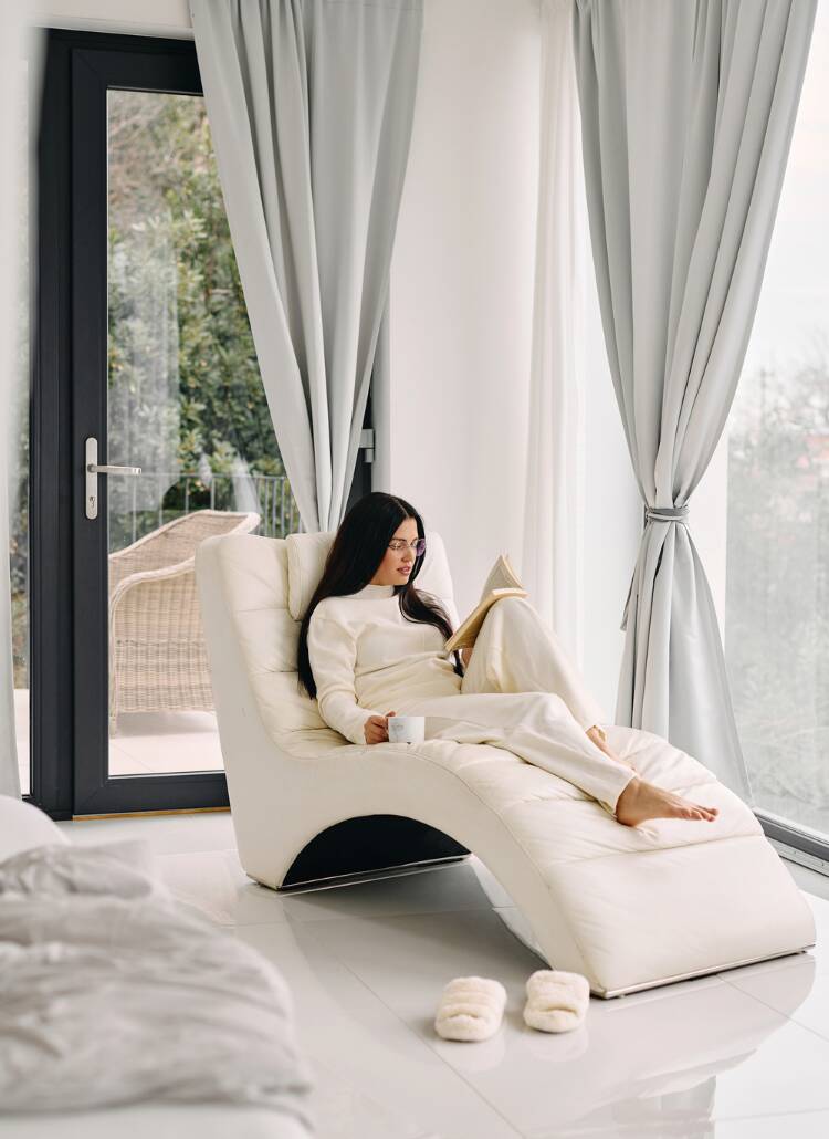 person relaxing on a white chaise lounge chair by large windows, reading a book and holding a coffee cup. They are wearing a cozy cream-colored sweater and matching loungewear, creating a serene monochromatic scene. A featured image in this blog post about Friday affirmations