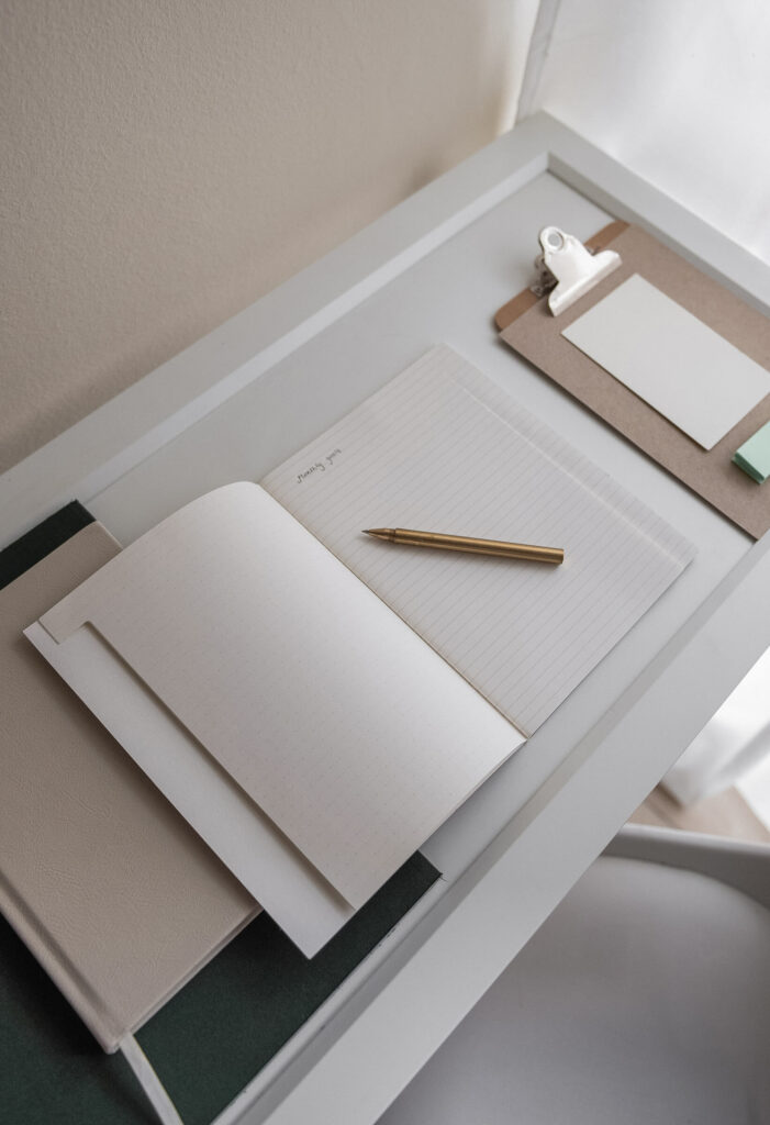 Minimal journaling setup with open notebook, gold pen, and clipboard on a white desk — aesthetic workspace for mindful planning and self-reflection.