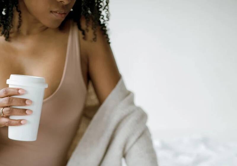 A woman with curly hair wearing a beige top and cardigan, holding a white takeaway coffee cup against a light background.