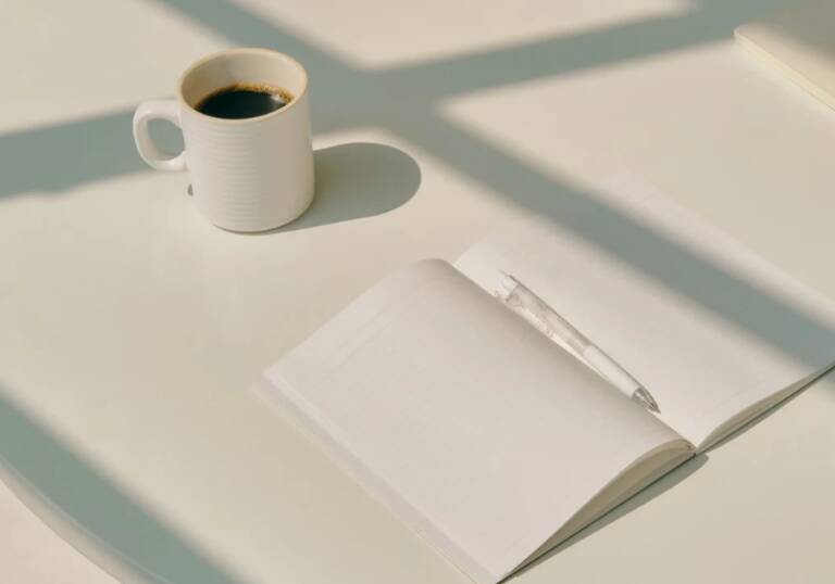 A white mug of black coffee beside an open, blank journal and pen on a sunlit desk, representing journaling and reflecting on life goals.