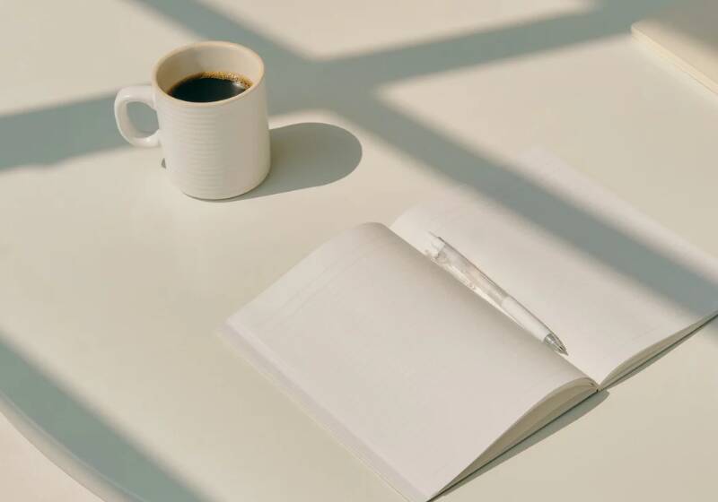 A white mug of black coffee beside an open, blank journal and pen on a sunlit desk, representing journaling and reflecting on life goals.