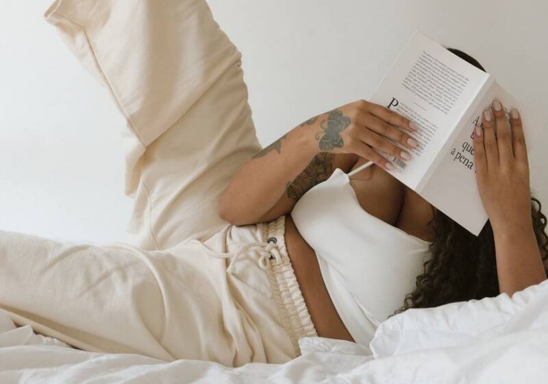 A woman with tattoos relaxing on a white bed in cream loungewear, reading a book with one leg raised against a minimalist white background.